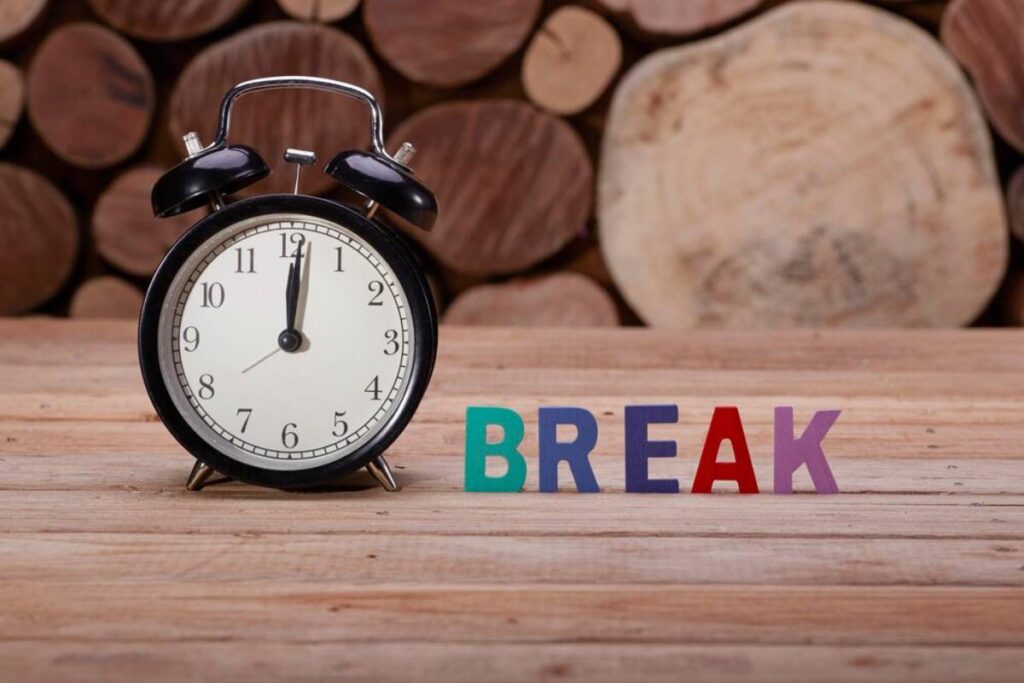 Black alarm clock showing 3 o'clock and colorful letters spelling BREAK on a wooden surface with logs in background.