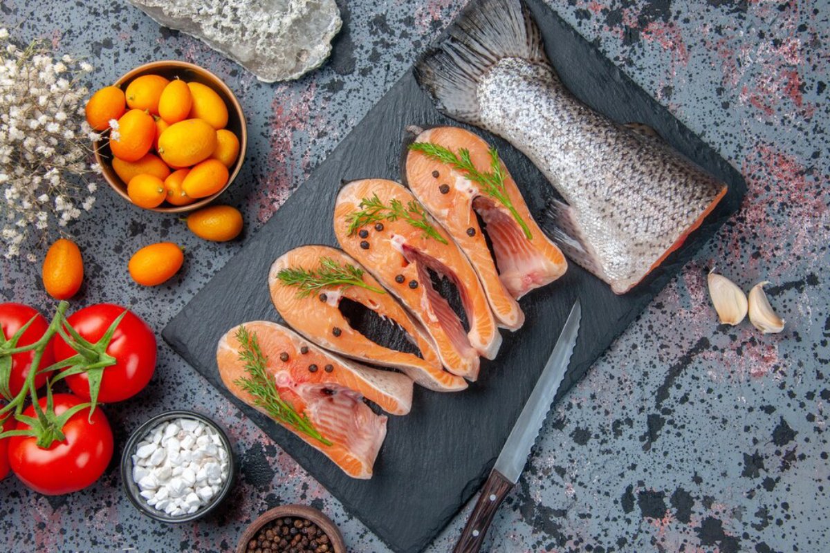 Fresh salmon steaks and whole fish on a slate with tomatoes, kumquats, and seasonings on a speckled table.