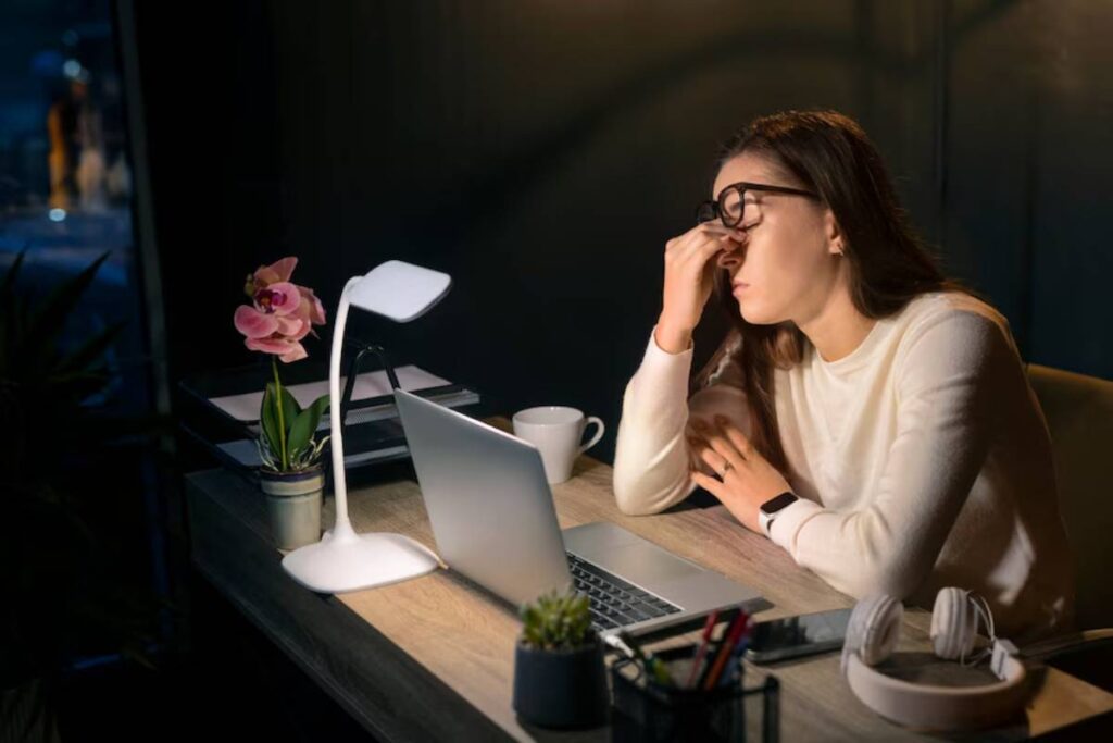 Woman in glasses looks tired while working late at night in a dark office lit by a desk lamp.