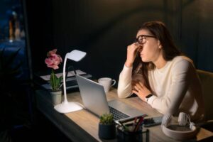 Woman in glasses looks tired while working late at night in a dark office lit by a desk lamp.