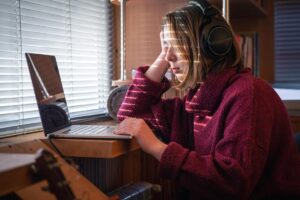 Young woman in headphones using a laptop by a window, looking thoughtful. Sunlight casts stripes across her face.