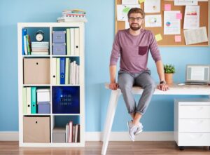 Young man sitting on a desk in a tidy home office with organized shelves and a blue wall.