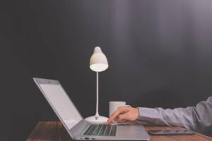 Person typing on a laptop under a lamp, with a dark background.