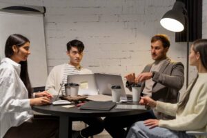 Four colleagues discuss work at a table with laptops and coffee in a modern office with a white brick wall backdrop.