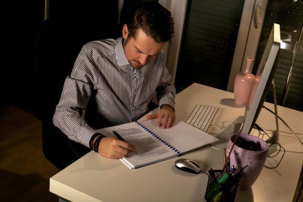 Man in striped shirt writing in notebook at a desk with computer and decorative items, in a dimly lit room.