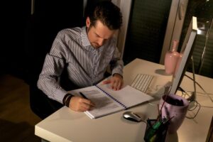 Man in striped shirt writing in notebook at a desk with computer and decorative items, in a dimly lit room.