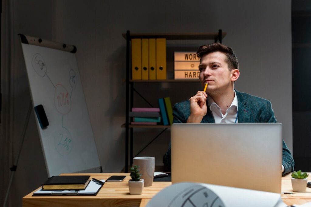 Man in suit pensively working at a laptop in a modern office, with a whiteboard and bookshelf in the background.