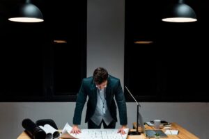 Man in suit examining documents at a desk under overhead lights in a dark office.