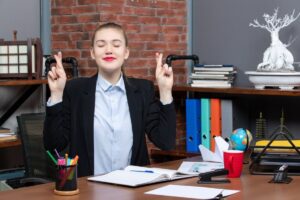 Young businesswoman at desk with eyes closed crossing fingers, expressing hope or making a wish.