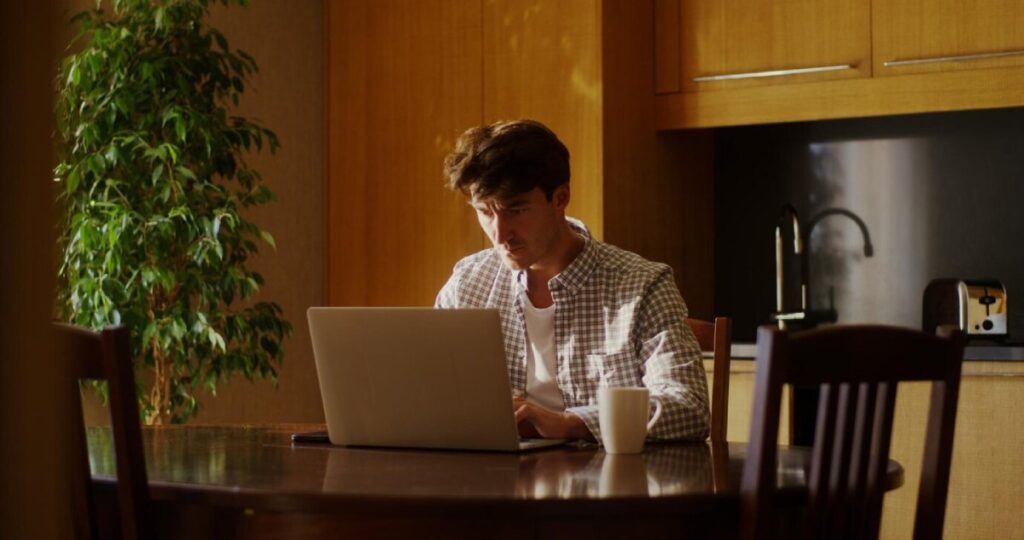 Man working on a laptop at a kitchen table, with a cup of coffee beside him.