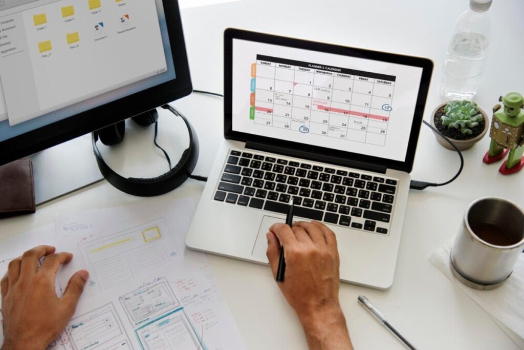 Person working on a laptop with a calendar on screen, surrounded by design sketches and a coffee cup.