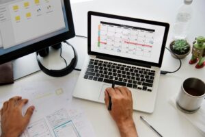 Person working on a laptop with a calendar on screen, surrounded by design sketches and a coffee cup.
