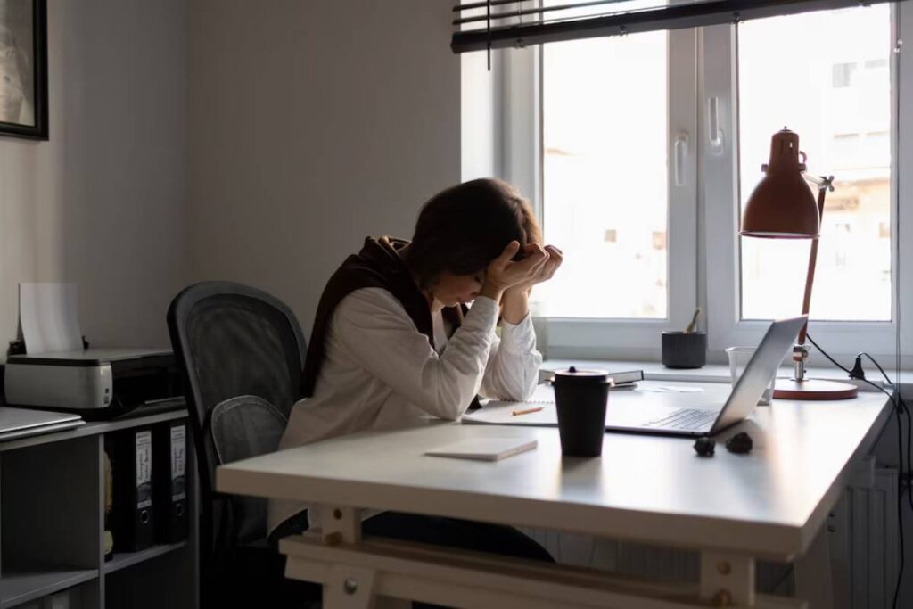 Woman sitting at desk, stressed, holding head over laptop in a home office.
