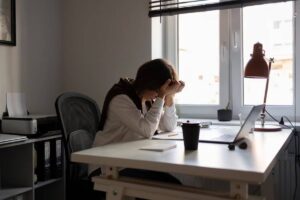 Woman sitting at desk, stressed, holding head over laptop in a home office.
