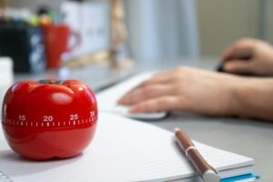 A tomato-shaped kitchen timer on a notebook, with a person working in the background.