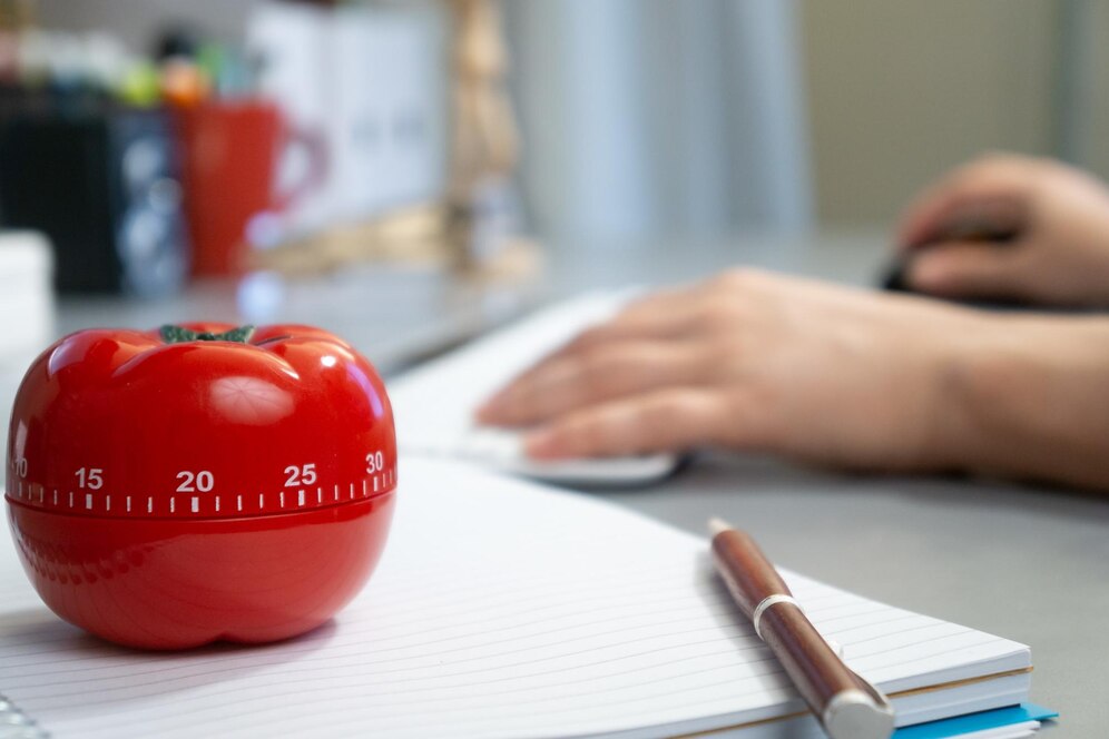 A tomato-shaped kitchen timer on a notebook, with a person working in the background.