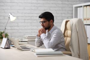 A focused individual sits at a desk with a laptop, books, and a lamp, deep in thought in a modern office setting.