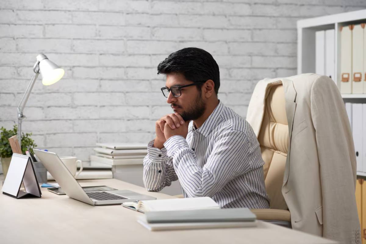 A focused individual sits at a desk with a laptop, books, and a lamp, deep in thought in a modern office setting.