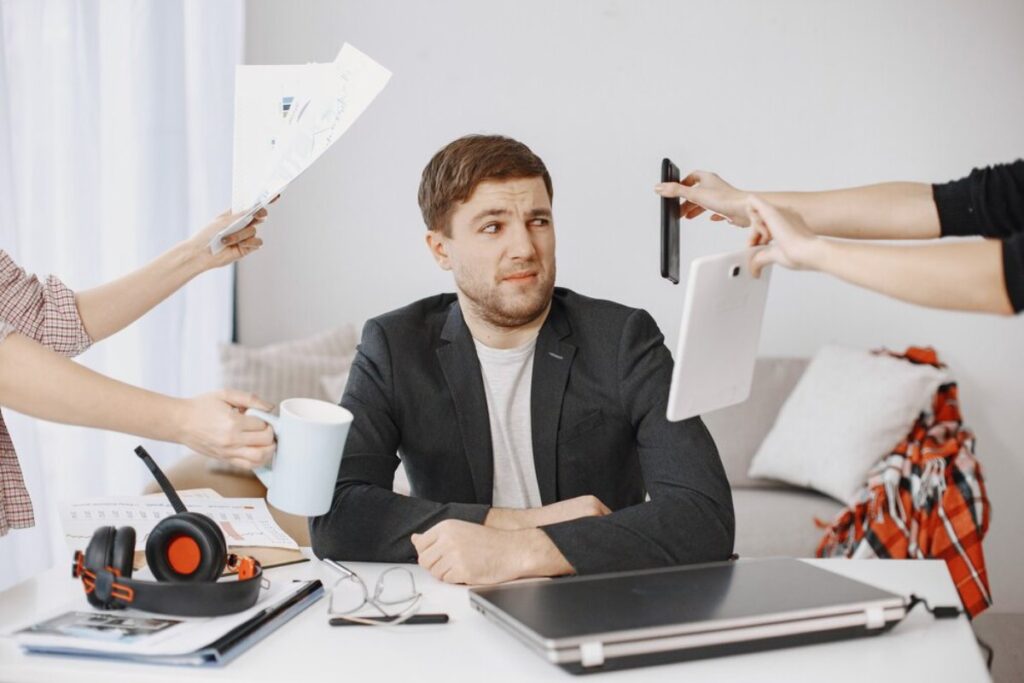 Man looking overwhelmed at a desk as hands hold a tablet, phone, and documents around him.