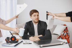 Man looking overwhelmed at a desk as hands hold a tablet, phone, and documents around him.