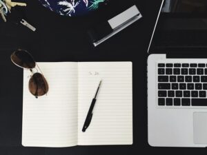 An overhead view of a desk with a laptop, a blank to-do list, a pen, sunglasses, and keys on a dark surface.