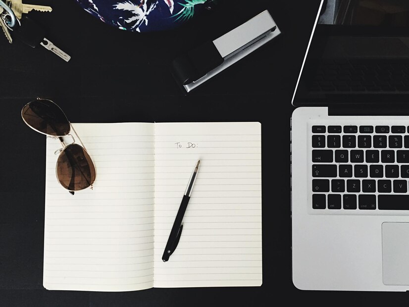 An overhead view of a desk with a laptop, a blank to-do list, a pen, sunglasses, and keys on a dark surface.