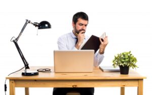 A man in a formal shirt sits at a wooden desk with a laptop, reading a book, accompanied by a desk lamp and a potted plant.
