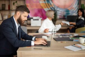 A man in a suit types on a laptop at a table filled with notebooks and materials, while two colleagues engage in discussion nearby.