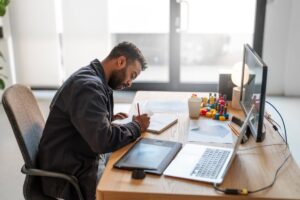 Man sketching on paper at his modern workspace with a digital tablet and laptop nearby.