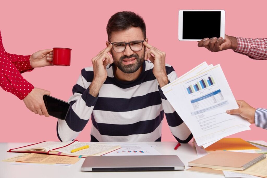 Stressed man at desk surrounded by hands holding various items like coffee, a phone, a tablet, and paperwork.