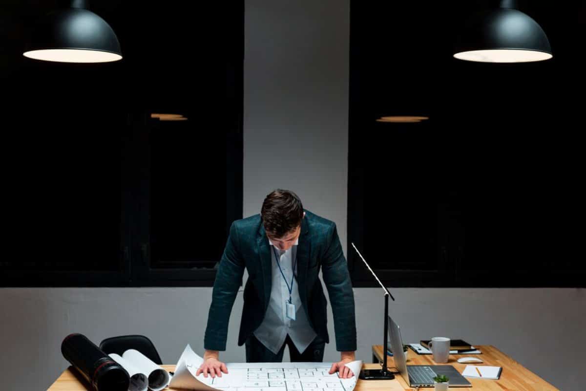 Man in suit examining documents at a desk under overhead lights in a dark office.